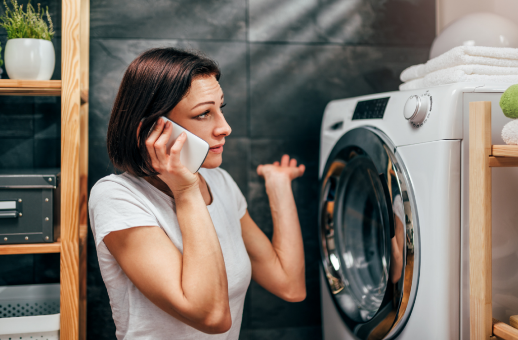 A woman converses on her cell phone while standing in front of a washing machine, engaged in a domestic setting.